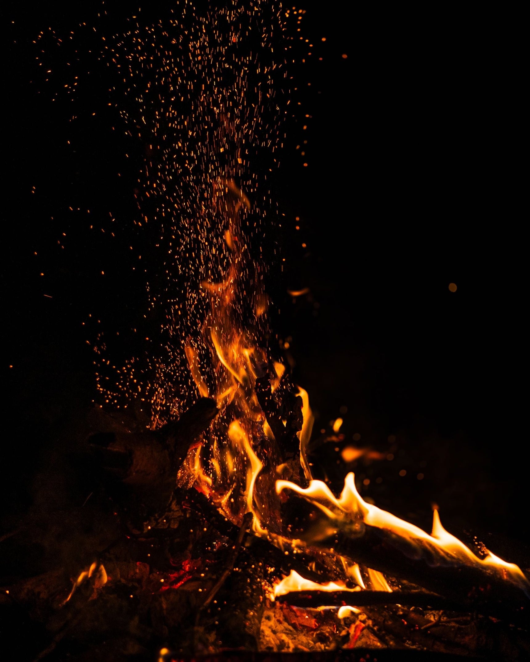 A close-up night scene of a campfire with bright orange and yellow flames rising from logs, with glowing embers floating upward against a black background.